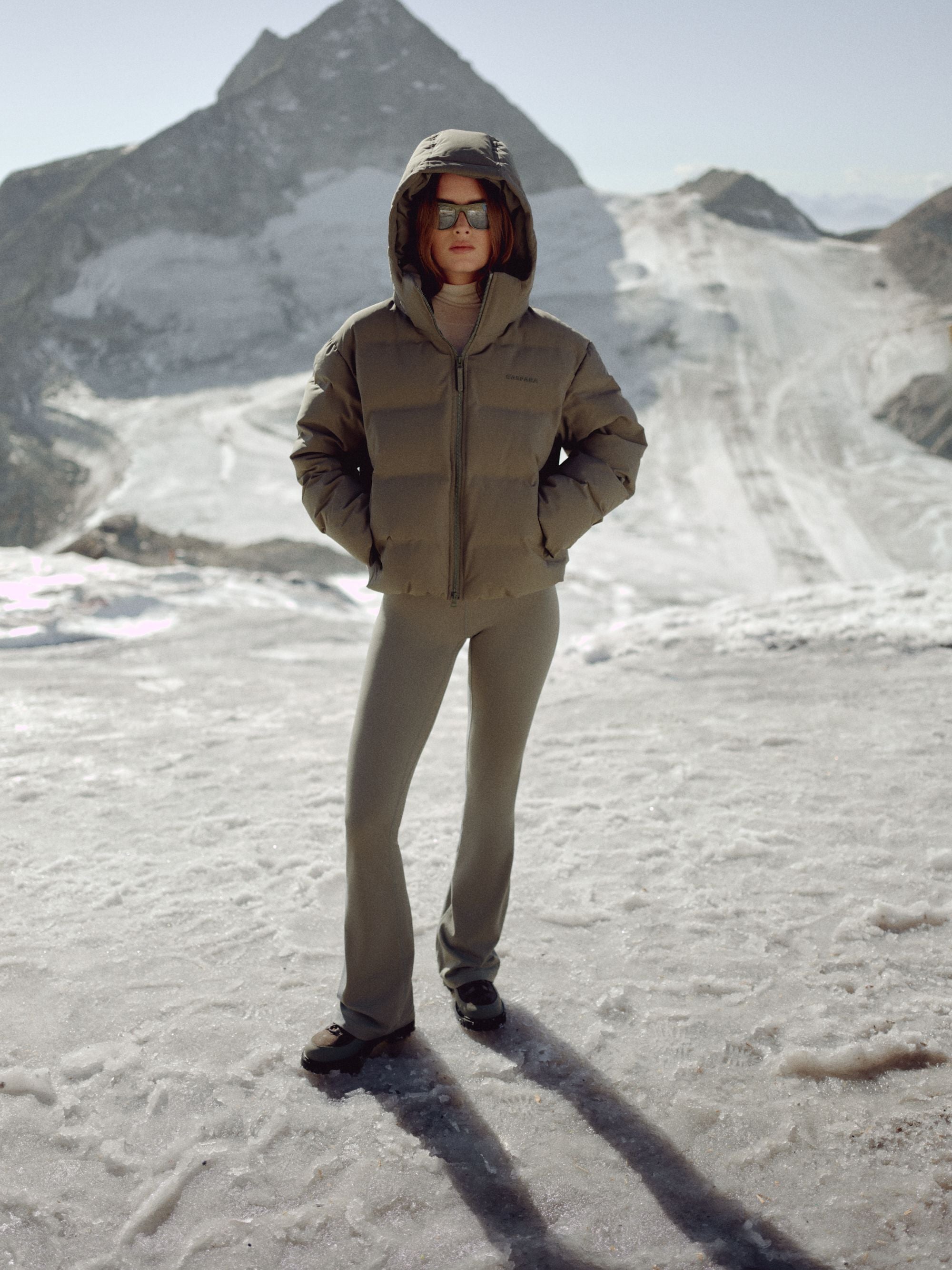 Woman wearing a khaki CASPARA Aurora puffer jacket with the hood up in a snowy mountain landscape.
